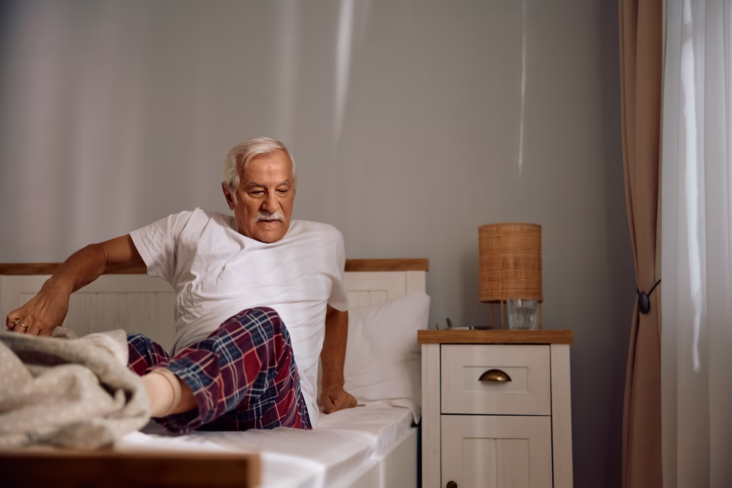 Wann mit Hüftschmerzen zum Orthopäden Älterer Mann steht morgens langsam aus dem Bett auf und stützt sich mit den Händen ab.