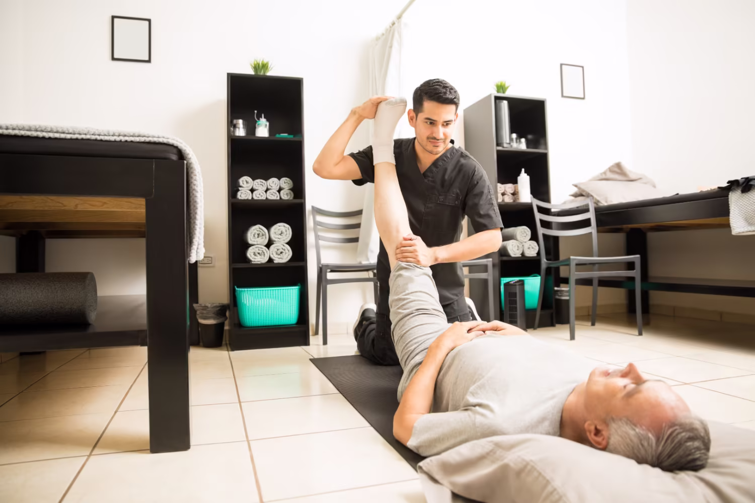 So behandeln wir Ihre Hüftarthrose Physiotherapeut dehnt das Bein eines älteren Patienten, der auf einer Matte liegt.