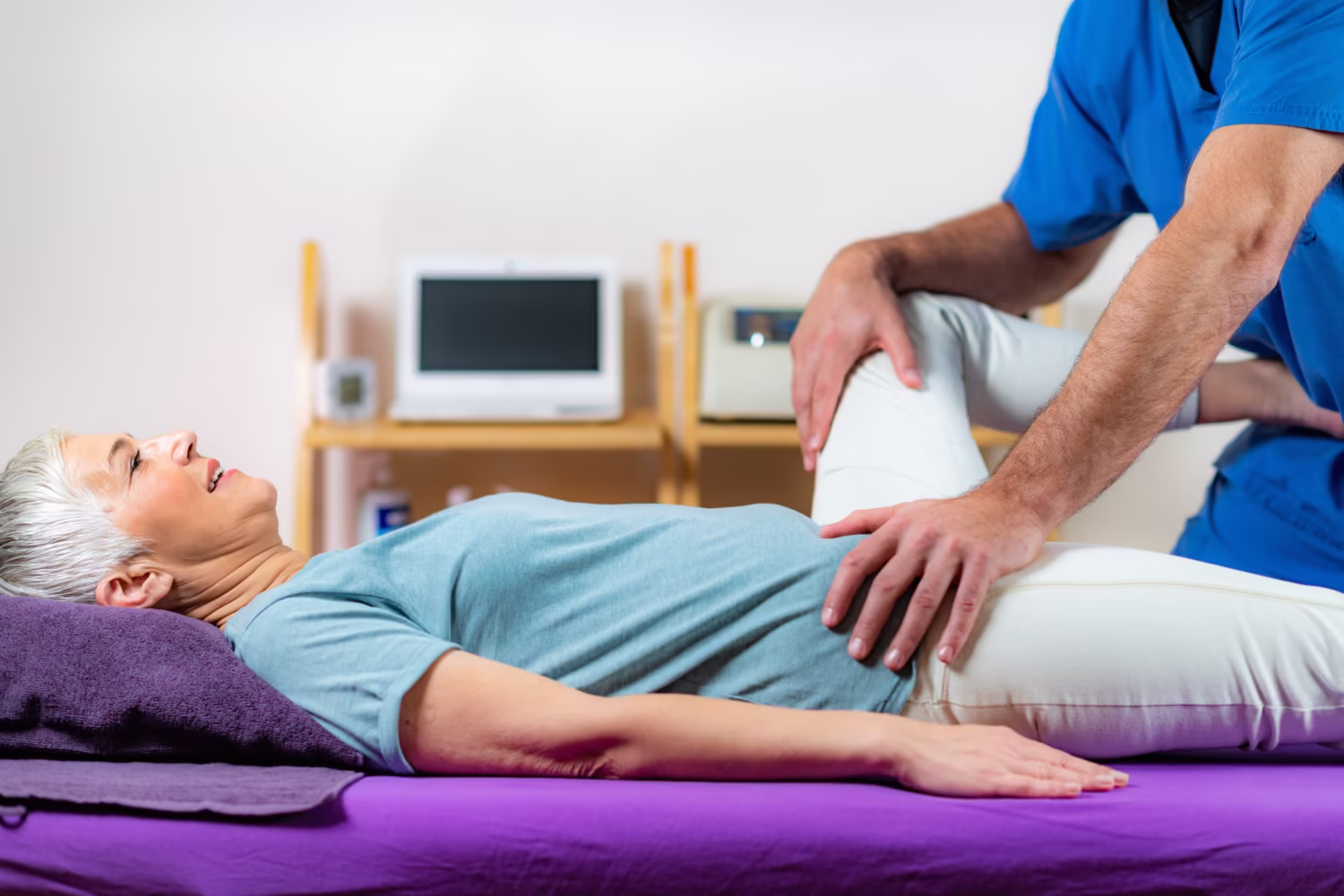 So untersuchen wir Ihre Hüfte Orthopäde untersucht mit beiden Händen die Hüfte einer älteren Patientin auf der Liege.